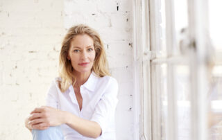 Close-up portrait shot of beautiful blond mature woman wearing white shirt and jeans while relaxing by the window. Ovarian cancer tested and clear